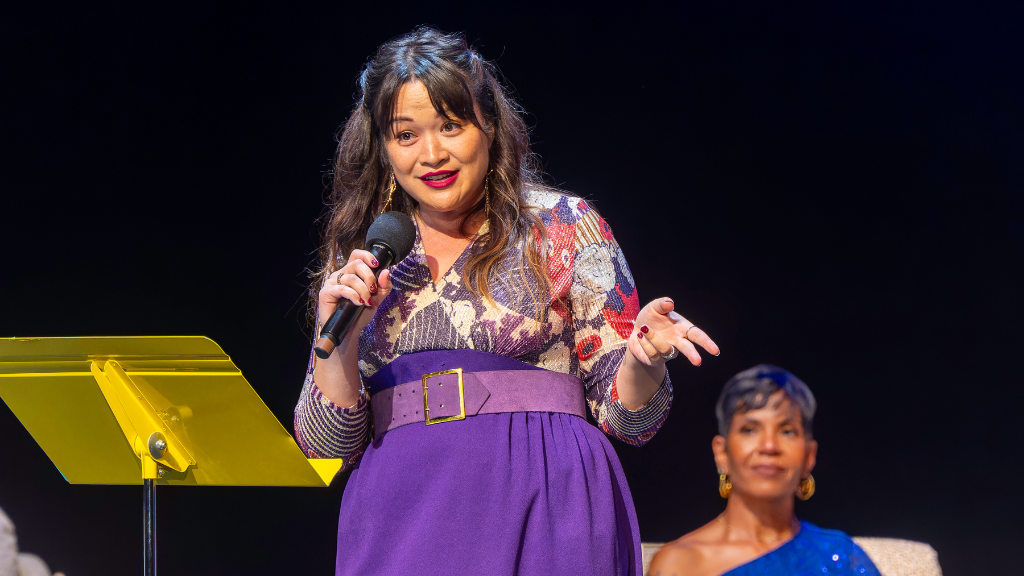 woman standing on stage behind a yellow stand holding a microphone