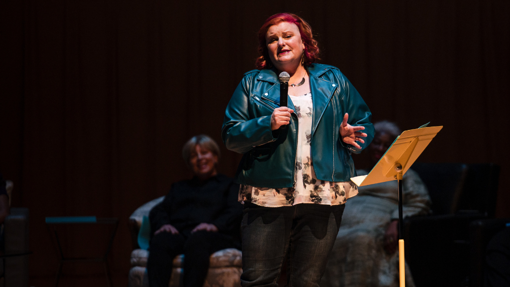 Woman stands on stage with microphone, other women sit behind her