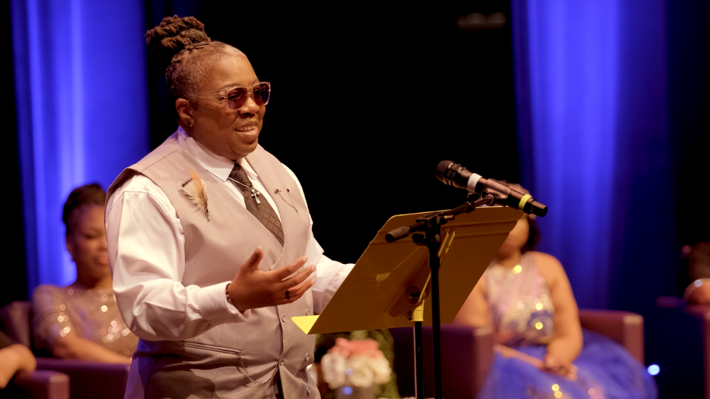 Woman stands on stage with microphone, other women sit behind her