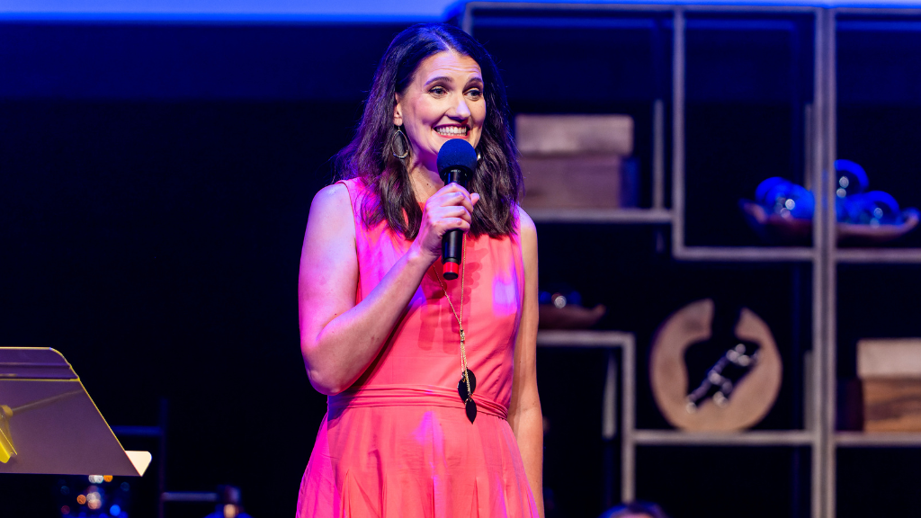 Woman holding a mic on stage, with another woman sitting behind her.