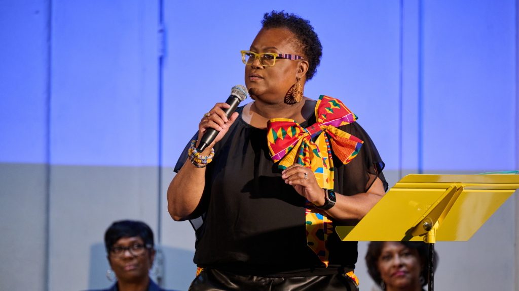 Woman stands on stage with microphone in right hand, two women sit behind her