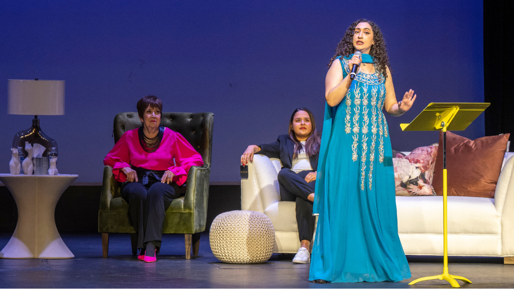 Woman stands on stage with microphone, other women sit behind her