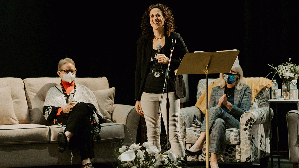 Woman holding a mic on stage laughing, women sit on couches behind her