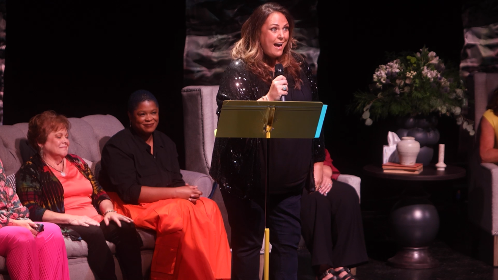 Woman stands on stage with microphone, other women sit behind her