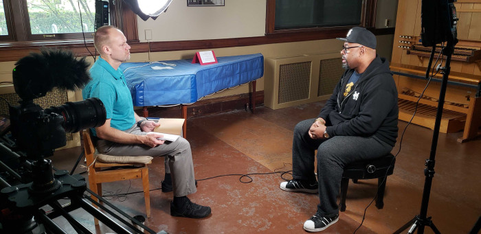 Two men sit down for an interview with cameras and lights