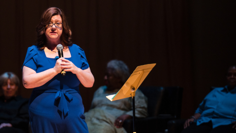 Woman stands on stage with microphone, other women sit behind her