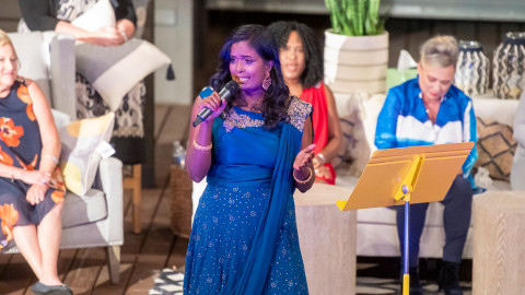 Woman stands on stage with microphone, other women sit behind her